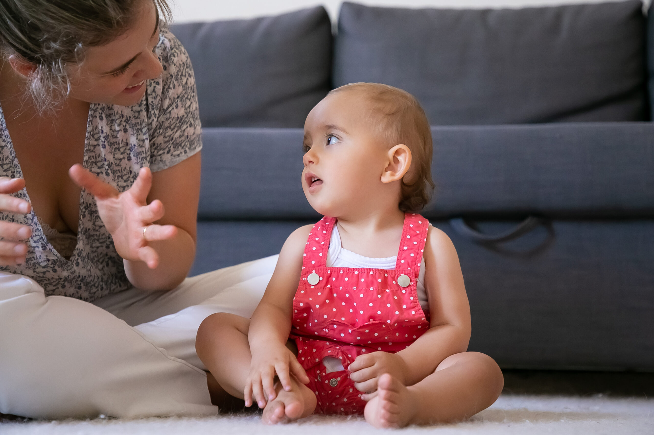 Cute little girl listening mom with open mouth and looking at her. Cropped mother sitting cross-legged on floor and talking to daughter. Lovely infant sitting barefoot. Weekend and motherhood concept