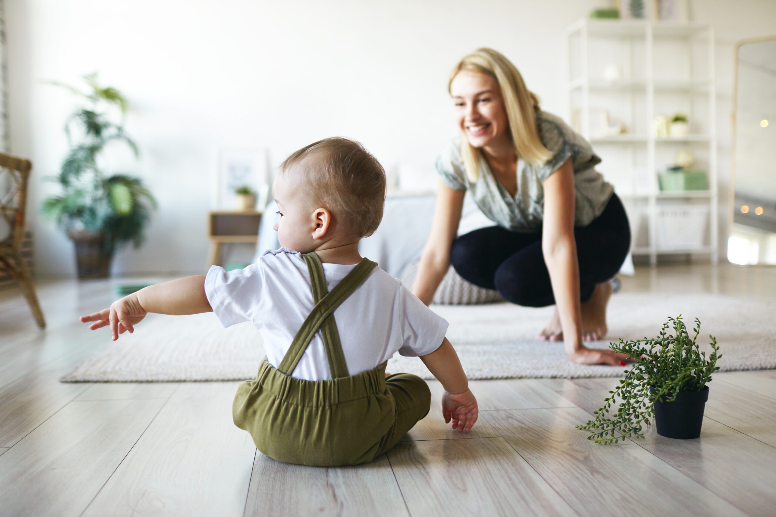 Back view of infant child in stylish clothes sitting on floor with plant pot, raising hand, pointing finger sideways, showing something to his young cheerful mother. Family, motherhood and infancy